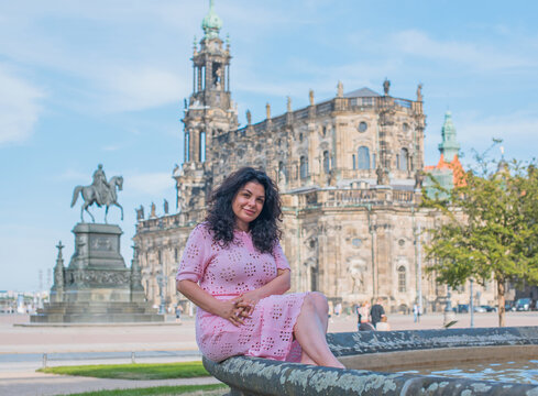 Beautiful brunette woman in pink dress sitting near fountain in Dresden, enjoying summer vacation with historic cathedral background in Saxony, Germany 