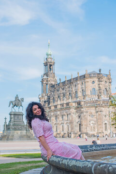 Beautiful brunette woman in pink dress sitting near fountain in Dresden, enjoying summer vacation with historic cathedral background in Saxony, Germany 