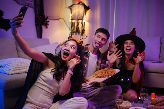 Young Asian friends taking a smartphone selfie with pizza at a Halloween party.