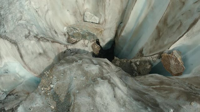 Overhead view of glacier ice cave showing textured ice walls, embedded rocks, deep blue ice. Natural lighting highlights cracks crevices. Evokes cold, remote, raw nature
