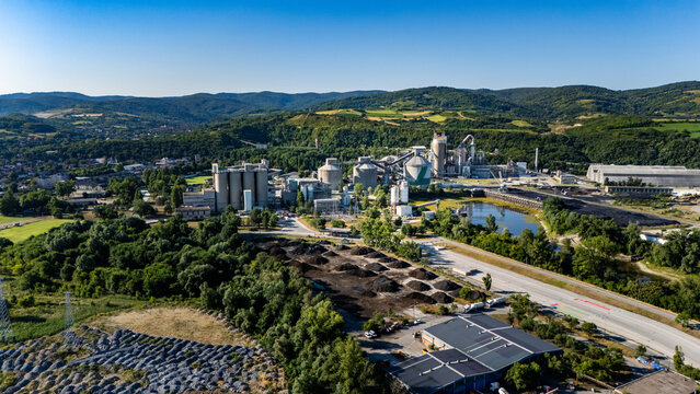24 July 2026: Aerial view of an industrial cement plant with large storage silos and processing facilities nestled in a green hilly landscape under a clear blue sky.