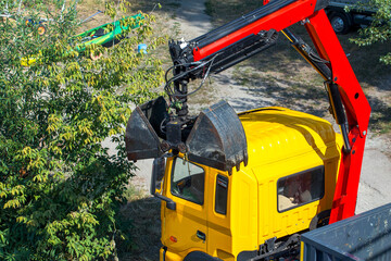 Elevated view of a yellow utility truck with a red hydraulic crane and black grapple attachment parked beside leafy trees on a sunny outdoor work site © ravital