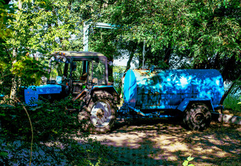 A blue farm tractor attached to a cylindrical water tank trailer stands in partial shade beneath leafy trees, creating a rustic rural transport scene © ravital