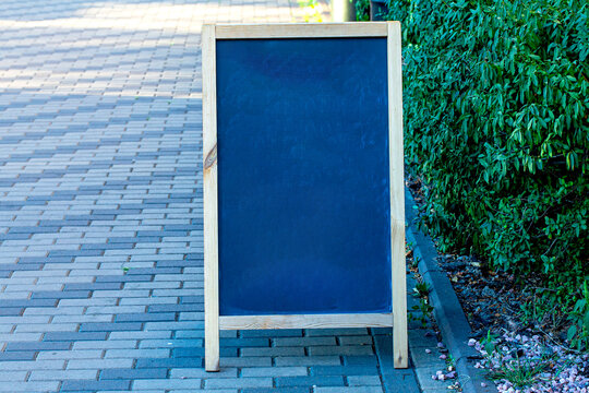 Empty black sandwich board with a natural wood frame placed on a paved sidewalk next to a green hedge, suitable for retail, cafe, restaurant, or street advertising