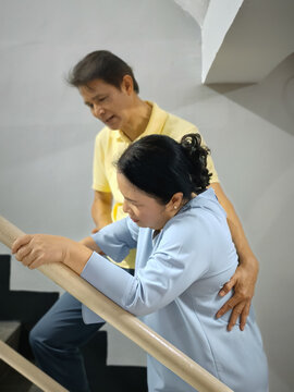 elderly asian man supporting a woman with knee joint pain or arthritis climbing staircase, holding her hand for stability and care
