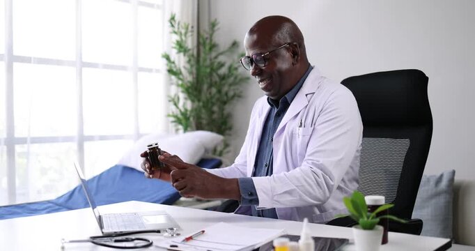 African american doctor holding a prescription bottle while typing on a laptop, providing telemedicine consultation or reviewing medication information in a medical office setting