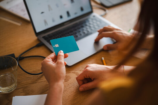A person holds a debit card while sitting next to someone using a laptop for online shopping. They are processing a digital payment at a wooden desk in an office.