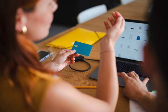 A woman holds a debit card while a man types on a laptop to shop online. They sit at a wooden desk collaborating on a purchase during their workday in a home office.