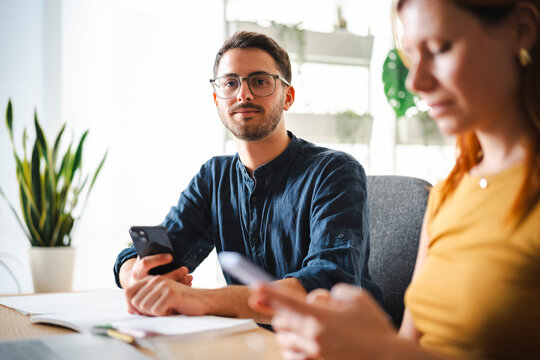 A young man wearing glasses sits at a desk with a blurred colleague in the foreground. They are both checking their smartphones while working in a bright office space.