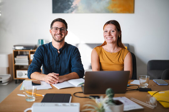 A man and woman work together from a comfortable home office desk. They look at the camera with friendly smiles while collaborating on a project with a laptop nearby.