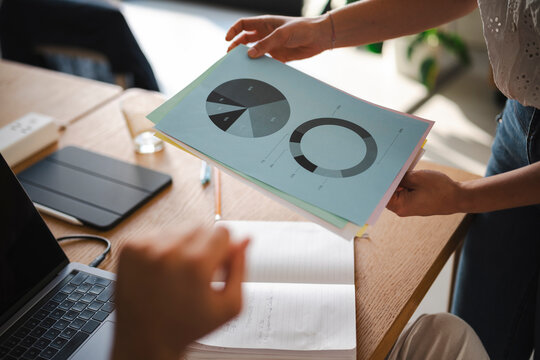 A professional presents business data and financial pie charts to a colleague during a collaborative meeting at a bright office desk with a laptop and notebook.