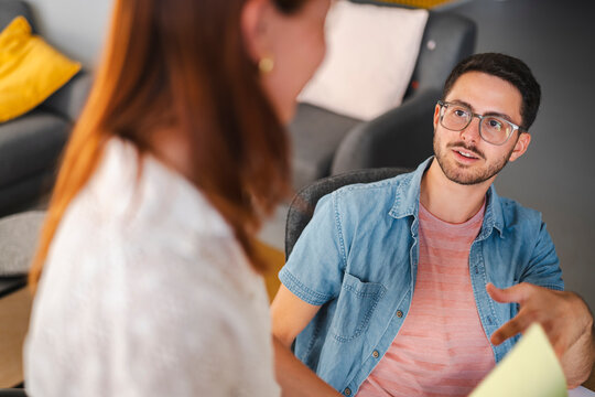 A professional man with glasses sits at his desk and gestures while talking to a female colleague. They are collaborating on a project in a modern office environment.