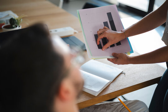 A professional colleague holds a chart and points to the data during a business meeting. They are collaborating on a project inside a bright and modern office space.