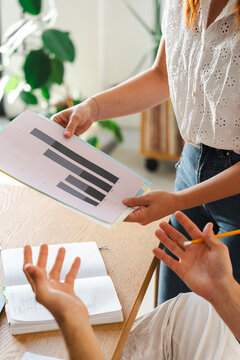 A woman shares a document showing business charts with a colleague at a wooden desk. They discuss the data and project goals in a bright, collaborative office space.