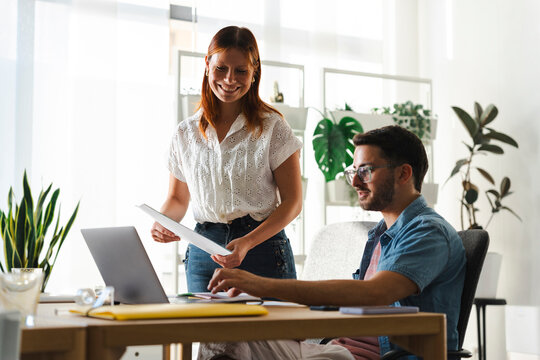 A man and a woman work together in a bright office. The woman holds documents while showing them to her colleague who is seated at a computer, discussing the project.