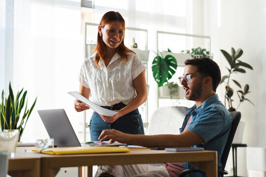 A woman stands next to her male colleague in a bright office. She holds papers while they review work on a laptop screen to discuss a project and solve tasks.