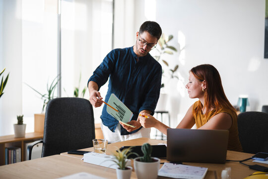 A man and a woman work together in a sunlit office, reviewing a printed graph. They collaborate on a business project while sitting at a wooden desk with laptops.