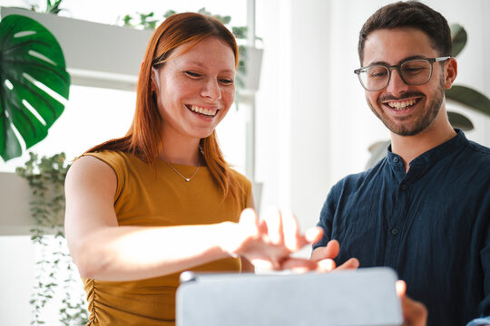 A young woman with auburn hair and a man in glasses laugh together while working on a digital tablet in a bright office filled with lush green indoor plants.
