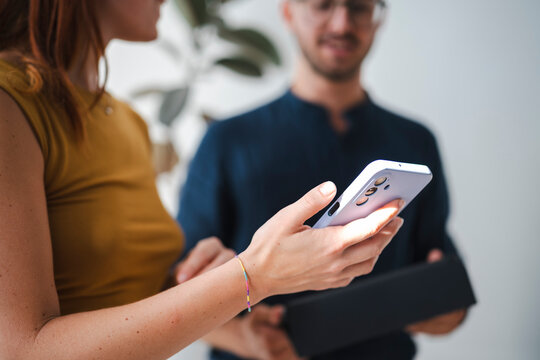 A young woman holds a new smartphone while a man stands behind her holding the product box. They are unboxing a modern gadget together in an indoor office space.