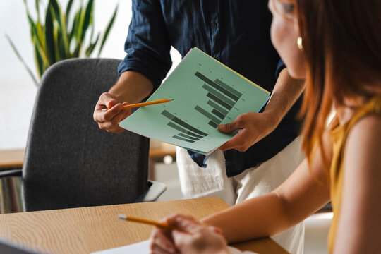 A business professional holds a green document with bar charts while explaining key data to a colleague in a modern office. They are collaborating on a team project today.