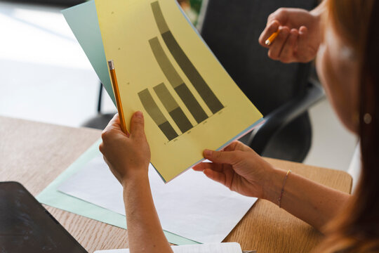 A professional holds a paper document featuring a detailed bar graph during an office meeting. A colleague points with a pencil to discuss the growth data results.