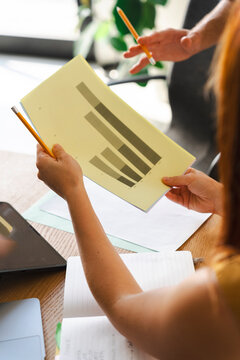 Two coworkers gather at a wooden desk to examine a printed bar graph. They use pencils to track data points and discuss progress during a collaborative meeting.