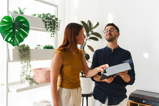 A man and a woman stand together in a brightly lit room filled with potted plants. The woman points to a tablet held by the man as they discuss a project together.