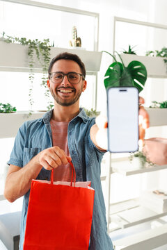 A young man wearing glasses holds a red shopping bag in one hand while displaying a smartphone with a blank white screen directly toward the camera in his home.