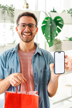 A happy young man with glasses holds a red shopping bag and displays a smartphone with a blank screen for app mockups. He stands in a bright room filled with plants.