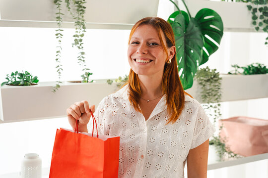 A young woman with red hair wearing a white shirt smiles at the camera while holding a bright red shopping bag. She stands in a room filled with green plants.