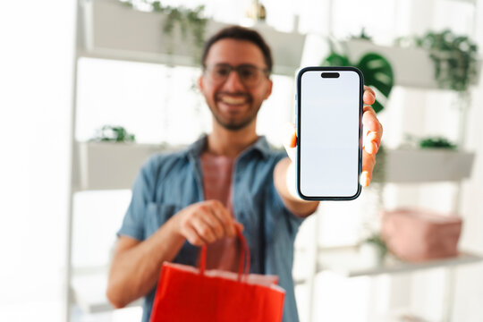 A happy young man holding a red shopping bag shows a smartphone with a blank white screen to the camera. This casual portrait highlights modern digital shopping.