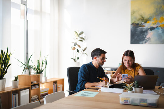 A male and a female colleague are collaborating in a bright modern office. They sit together at a desk, reviewing data on a smartphone while referencing a laptop.