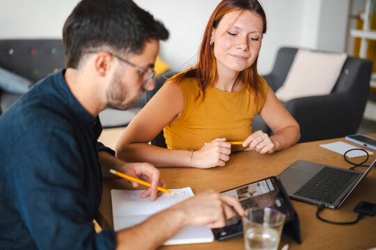 A professional man and woman sit together at a wooden desk to collaborate on a digital project. They use a tablet and laptop to share ideas while writing in a notebook.