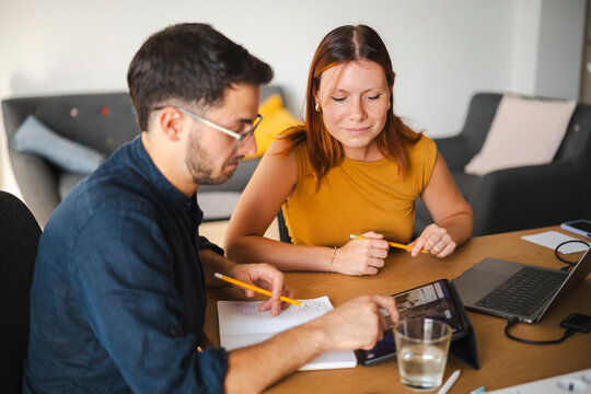 A young man and woman sit together at a desk working on a collaborative project. They use a tablet and laptop to research while taking notes in a notebook at home.