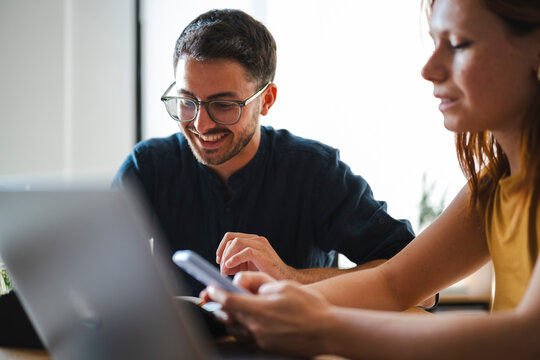 A young man and woman are collaborating in a bright modern workspace. They work together while reviewing content on a laptop screen and checking a mobile device.
