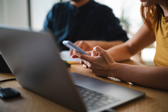 A woman holds a smartphone while sitting at a wooden desk with a laptop. A colleague works on notes in the background during their collaborative workspace session.