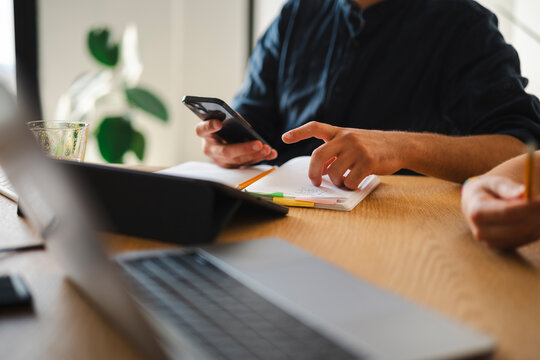 A person in a dark shirt sits at a wooden table, checking their smartphone while reviewing handwritten notes in an open notebook. A laptop and tablet are nearby.