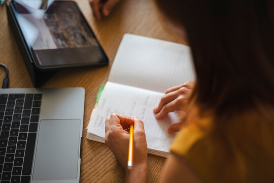 A woman sits at a wooden desk while writing notes in a physical notebook with a yellow pencil. A laptop and a digital tablet are placed nearby on the table surface.