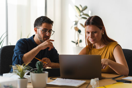 A man and a woman sit together at a desk in a modern office space. They are collaborating on a digital project, pointing at the laptop screen to discuss the work.