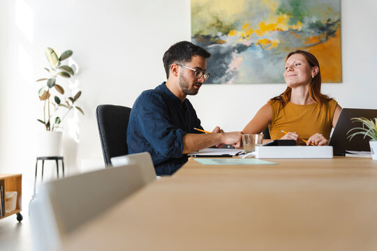 A man and a woman are working together at a desk in a bright home office. They discuss project notes on a laptop while collaborating on a professional task.