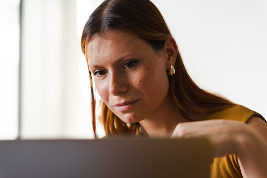 A focused young woman with reddish hair works diligently on her laptop computer. She is sitting in a brightly lit room and studying content on her screen intently.