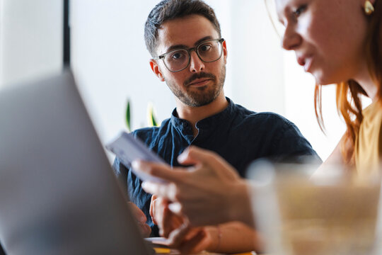 A young man with glasses and a woman collaborate while working together at a desk. They focus their attention on a laptop screen and a mobile phone during a meeting.