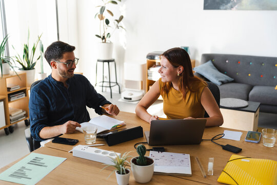 A young man and a woman work together in a bright home office, sharing ideas and taking notes while sitting at a wooden desk with a laptop and documents during the day.