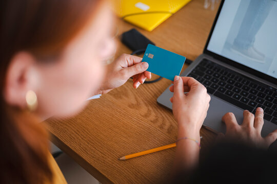 A woman sits at a wooden desk with a laptop while holding a debit card. She is entering payment information online to complete a transaction for her purchases.