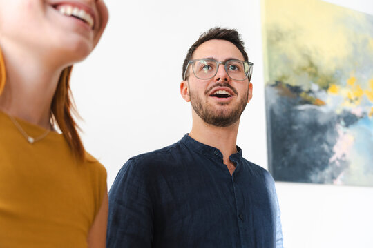 A young man wearing glasses and a woman with red hair stand in an art gallery, looking at a vibrant abstract painting with expressions of genuine wonder and interest.
