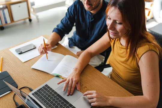 A young man and woman sit at a wooden desk collaborating on a project. They review information on a laptop while taking notes in a notebook during their workday.