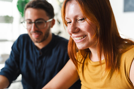 A young professional woman with red hair and her male colleague sit together in a bright office space. They are smiling while collaborating on a shared project.