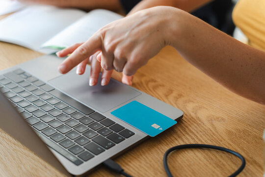 A person uses a laptop to manage finances online. Their hand navigates the trackpad next to a blue bank card placed on the keyboard surface during a remote work session.