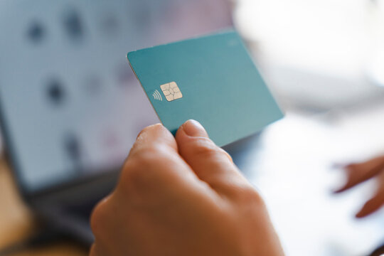 A person is holding a plastic credit card in their hand while preparing to make an online payment. They are sitting in front of a laptop computer to shop securely.