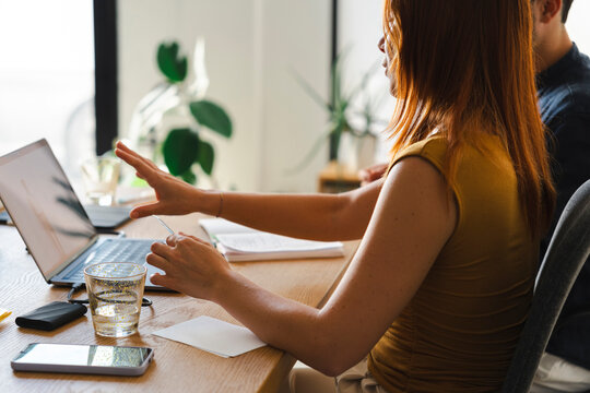 A woman with auburn hair sits at a desk while holding a small card and gesturing toward a laptop screen. She is collaborating with a colleague in a home office.